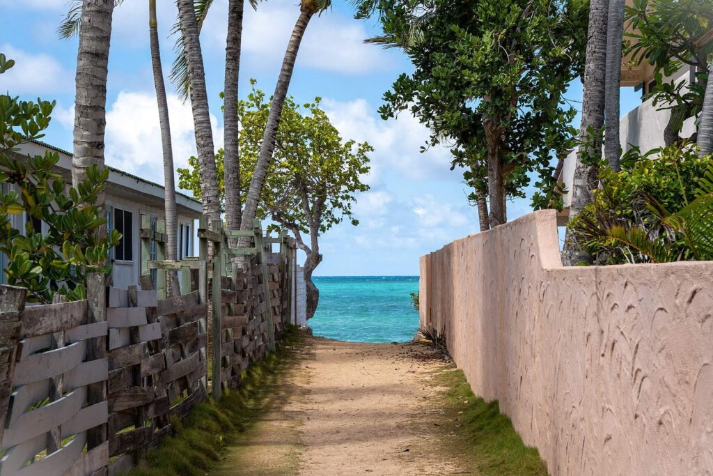A sandy public beach access path between a wooden fence and a pink wall, opening onto turquoise water at Lanikai