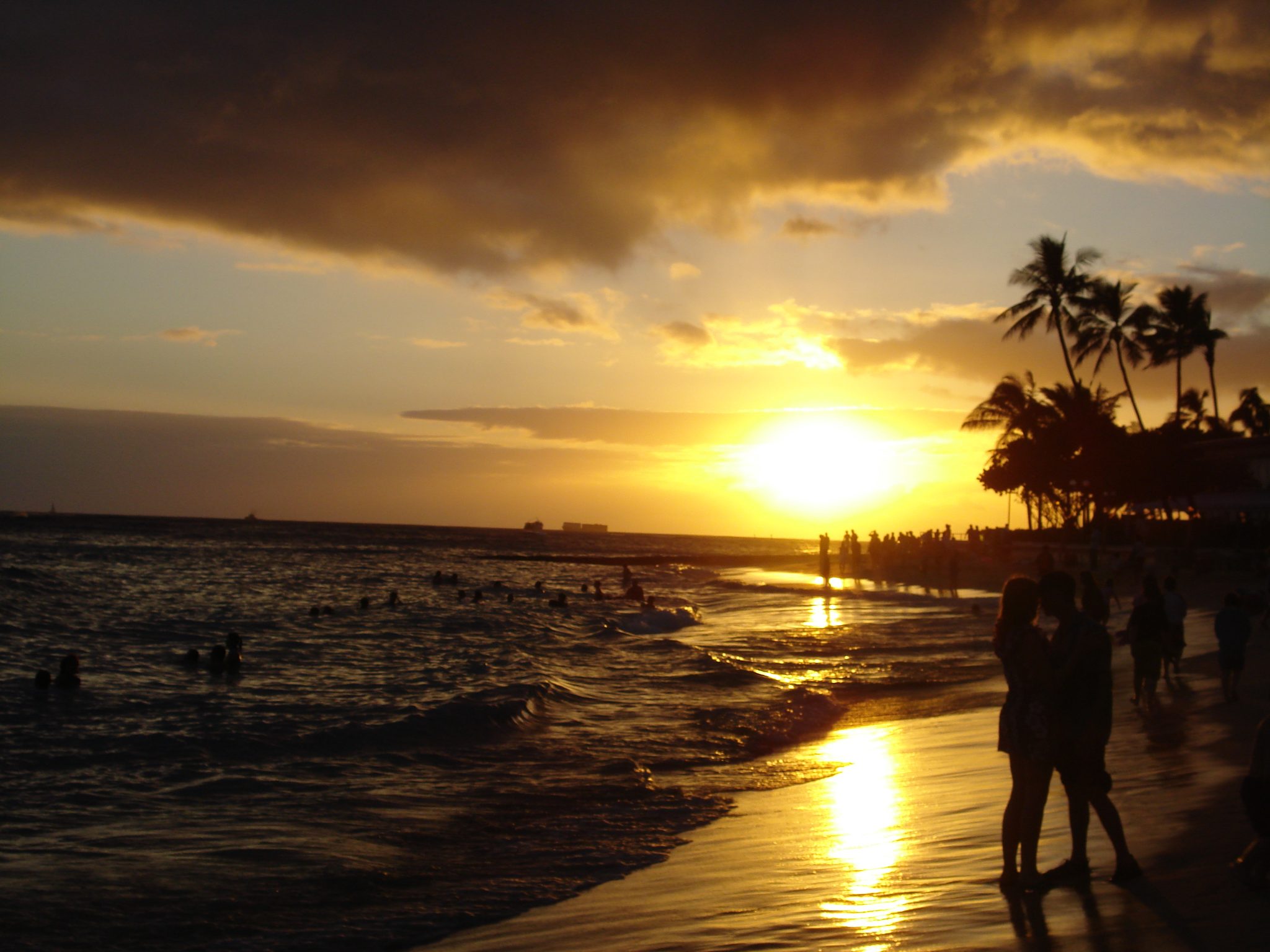The sun setting over the ocean at Waikiki Beach, with palms and people on the sand in silhouette
