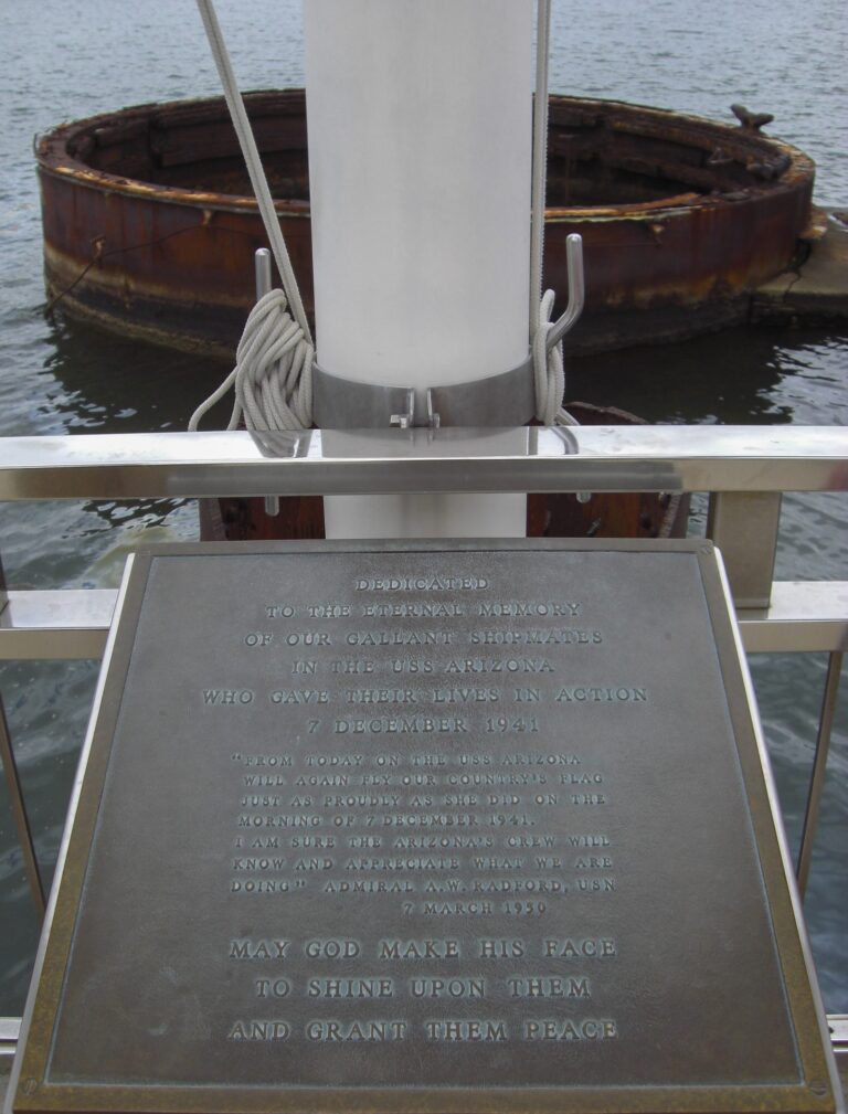 The dedication plaque at the USS Arizona Memorial flagpole, with the rusted wreck of the battleship beyond the rail