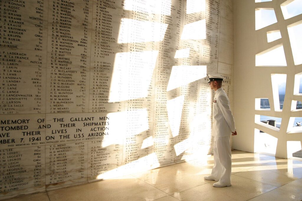 A Navy sailor in dress whites stands before the marble wall of names in the shrine room of the USS Arizona Memorial