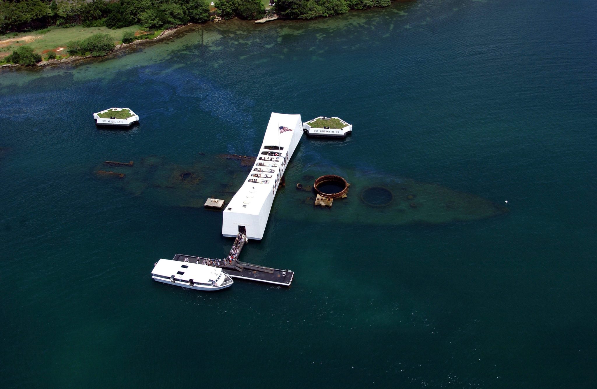 Aerial view of the USS Arizona Memorial at Pearl Harbor, with a Navy shuttle boat at the landing and the outline of the sunken battleship beneath the water