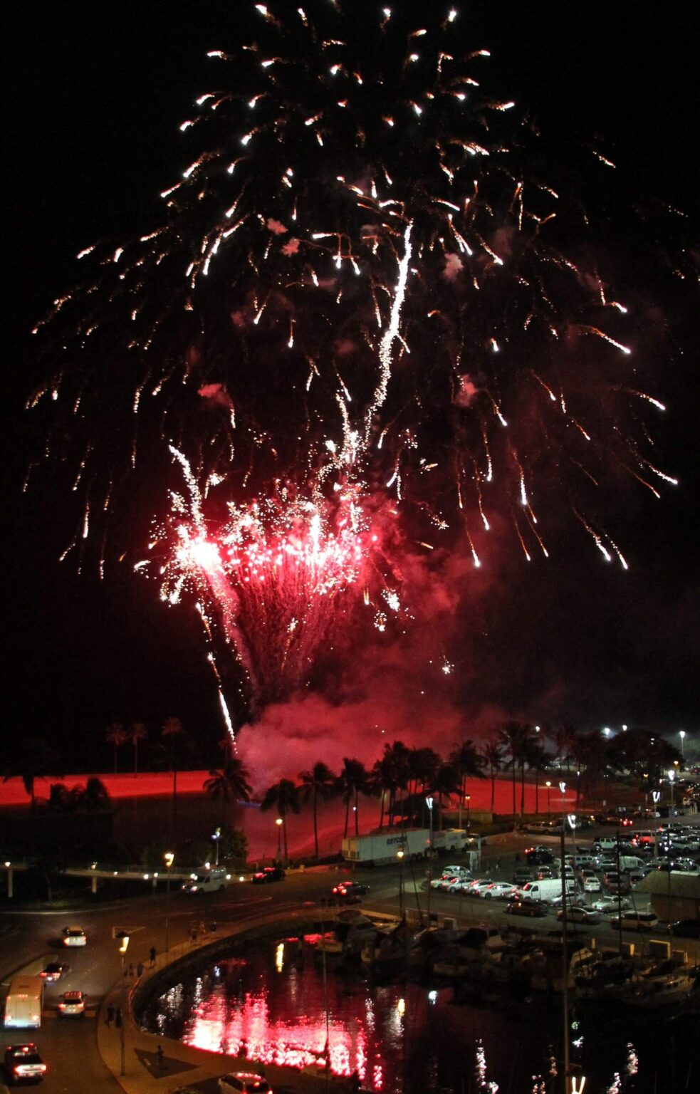 Red fireworks bursting over Waikiki at night, above a lit boat harbor