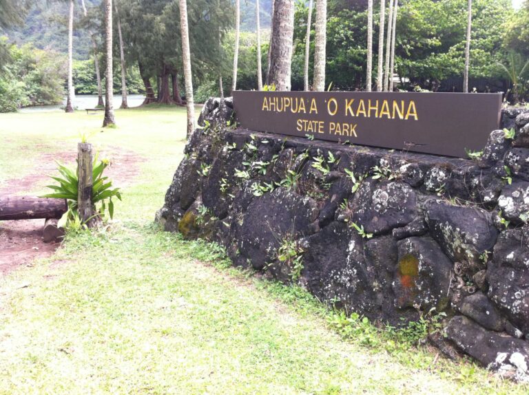 The Ahupuaʻa ʻO Kahana State Park sign on a lava-rock wall beside a lawn and palms