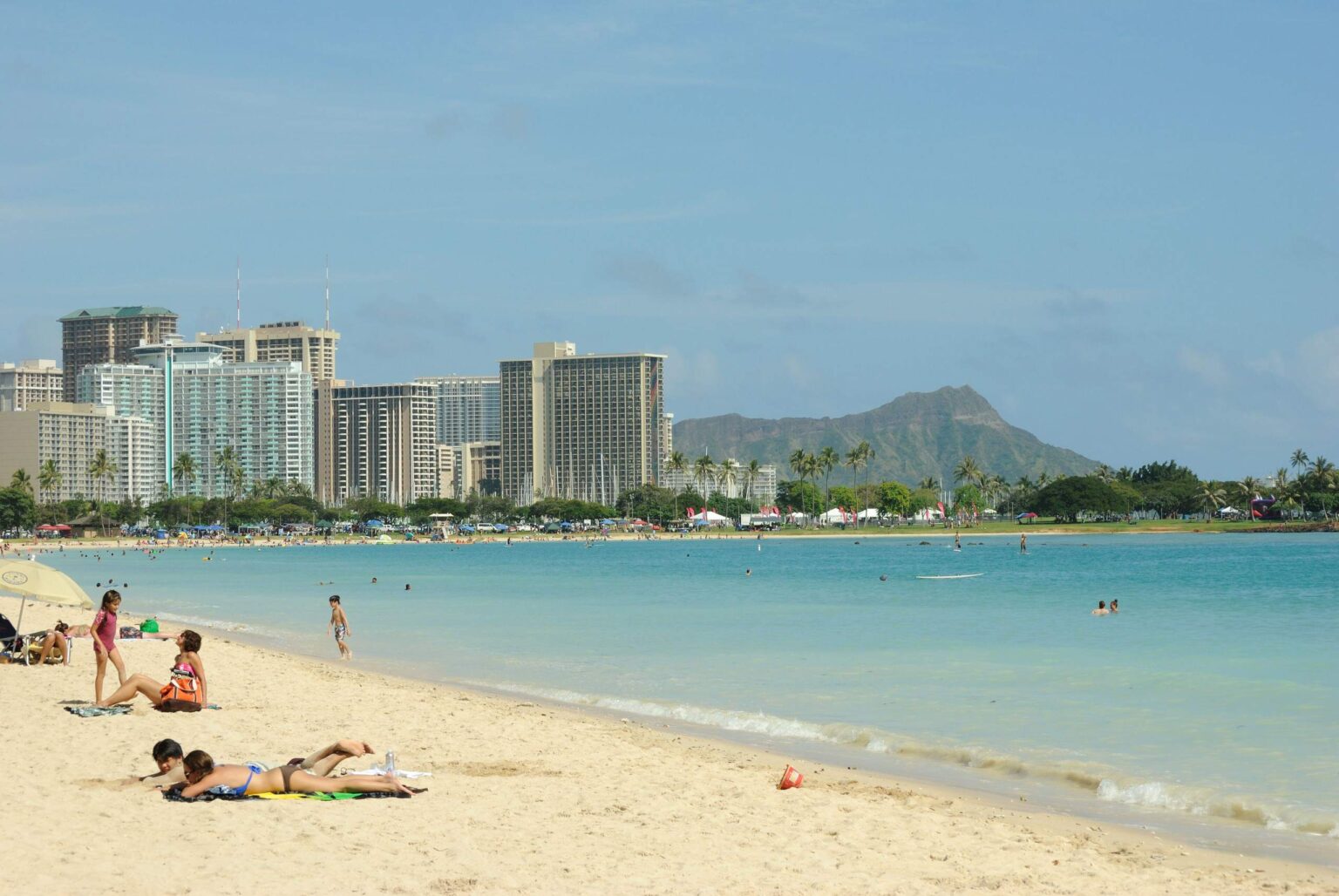 Families on the sand at Ala Moana Beach Park beside the calm swim channel, with Waikiki and Diamond Head behind