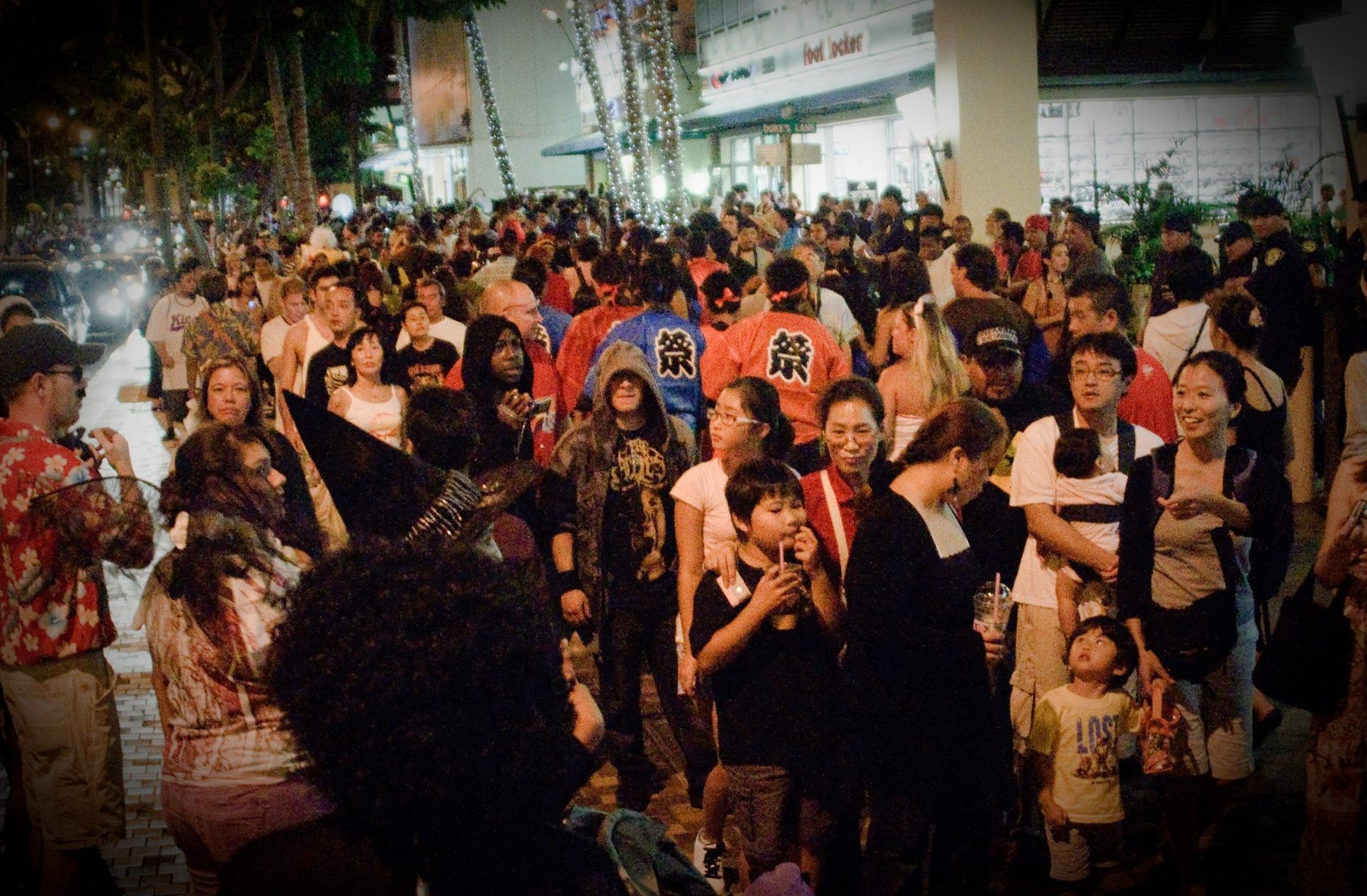 A crowd in Halloween costumes fills a Waikiki sidewalk at night, with families and kids among them