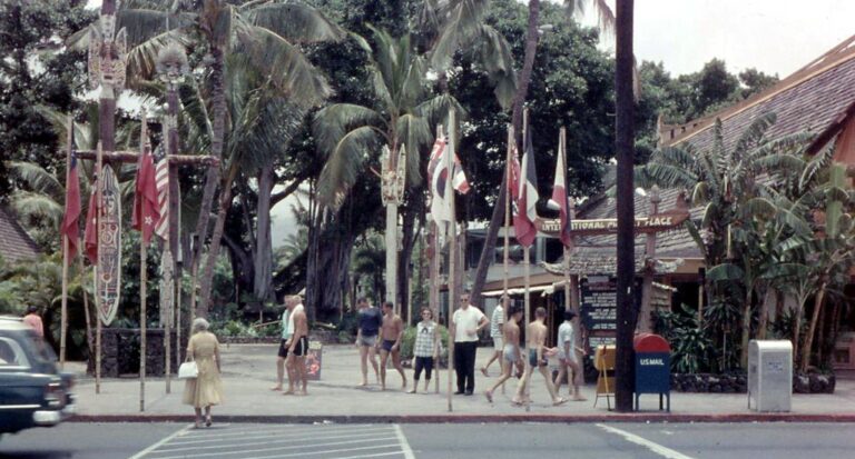 Flags and carved tiki poles at the entrance of the International Marketplace in Honolulu in 1958, with people crossing the street