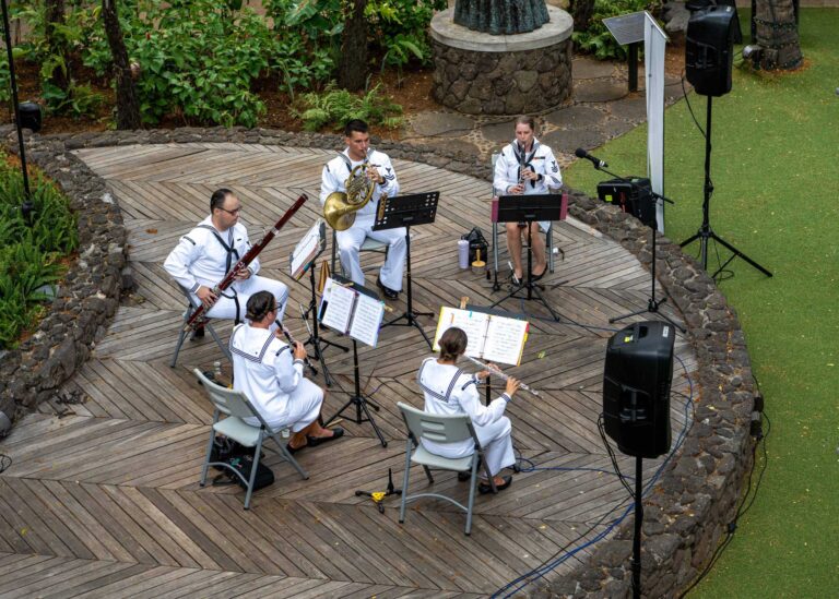 A U.S. Navy woodwind quintet in white uniforms playing on a wooden stage ringed by lava rock at International Market Place