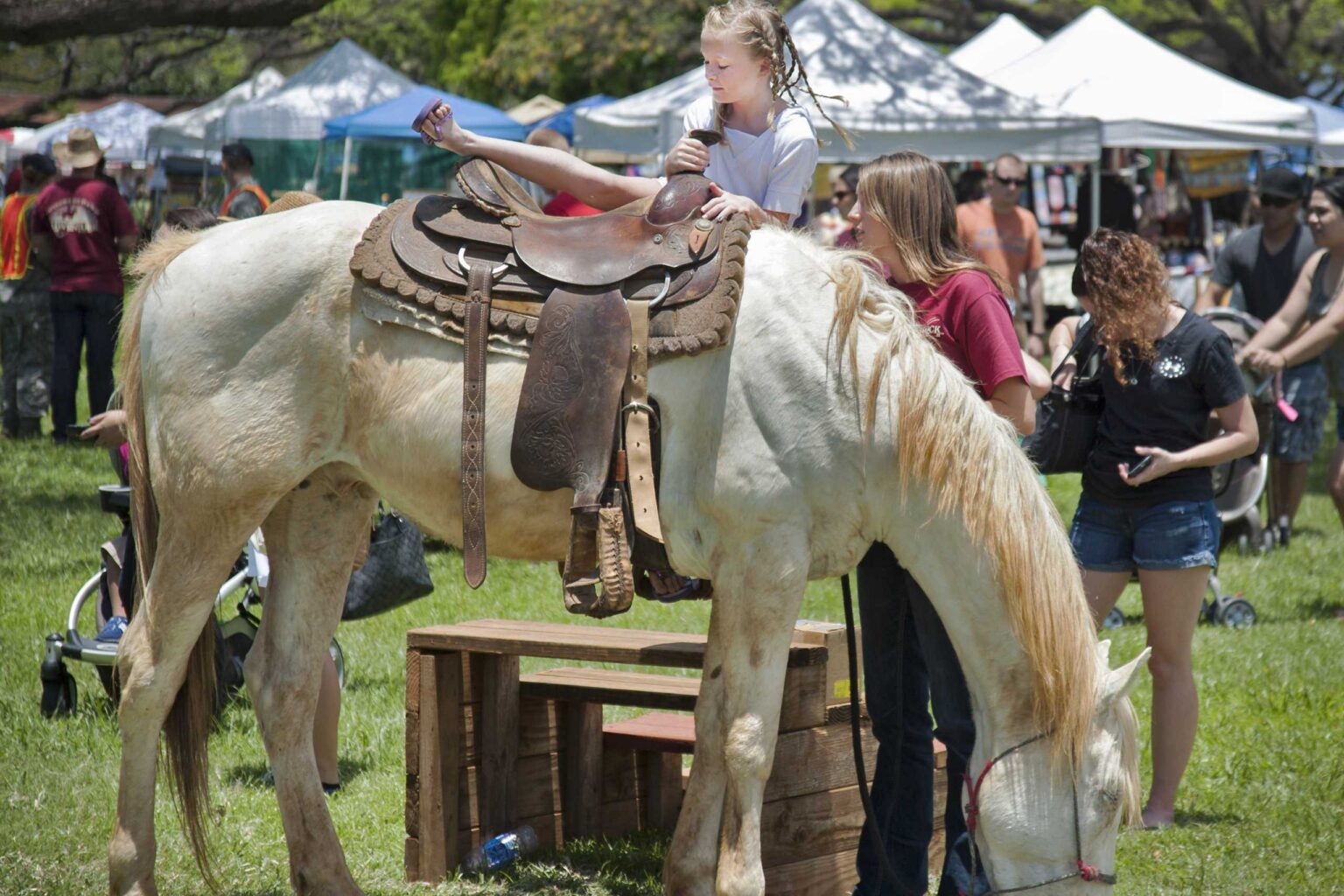 Shoppers browsing white and blue vendor tents on a grassy field at a craft fair on Oahu, with a child on a horse in the foreground