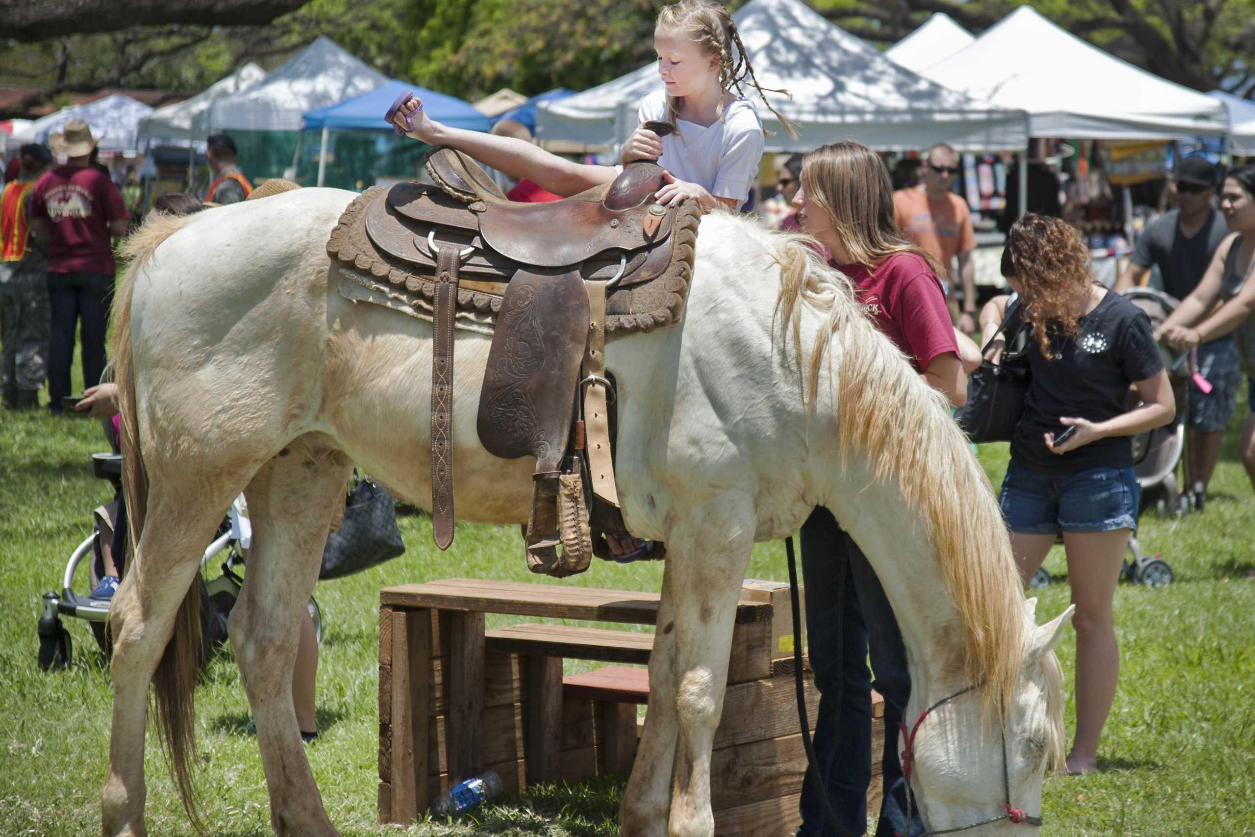 Shoppers browsing white and blue vendor tents on a grassy field at a craft fair on Oahu, with a child on a horse in the foreground