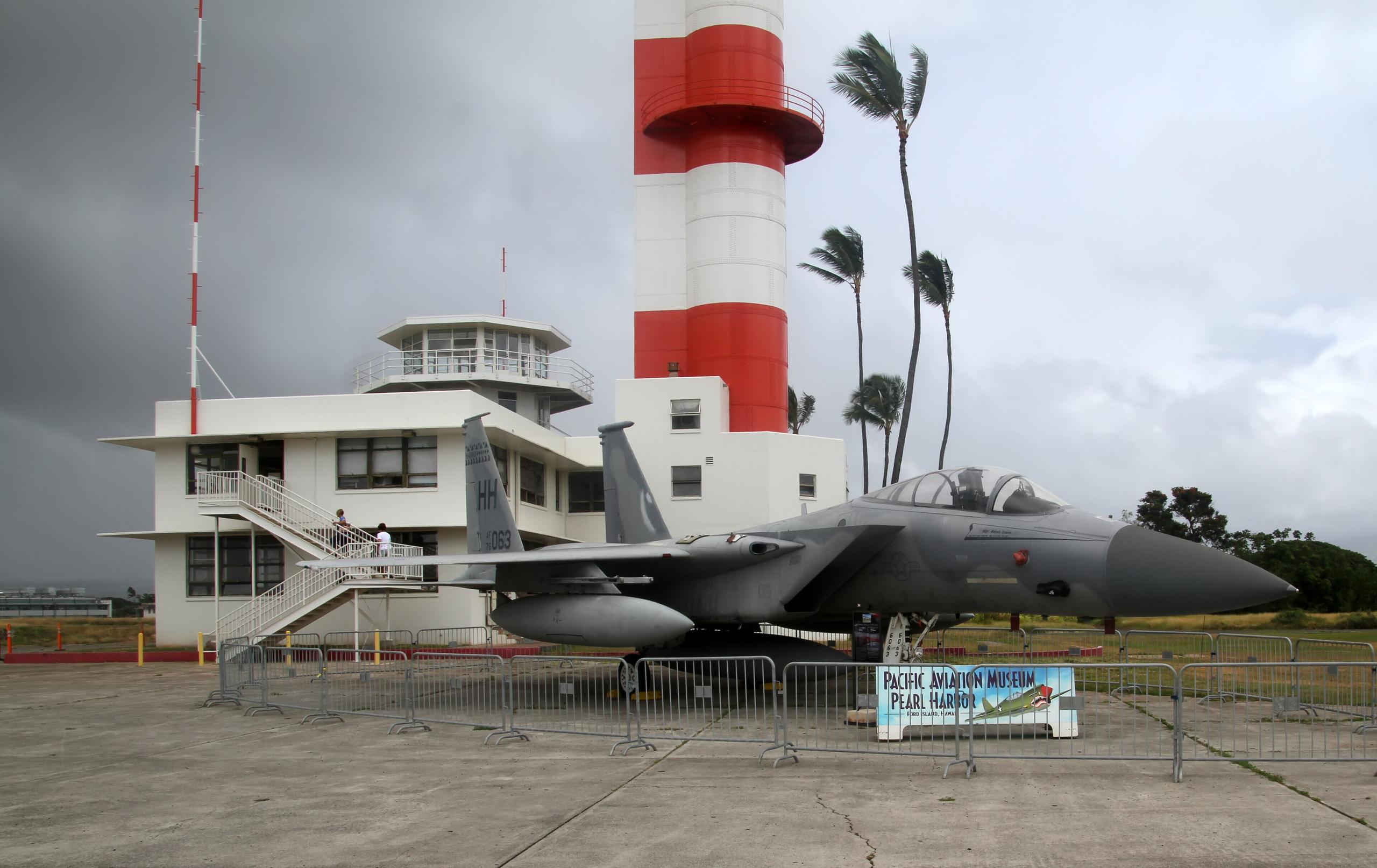 The red and white Ford Island Control Tower and the Operations Building at the Pearl Harbor Aviation Museum, with an F-15 on display in front