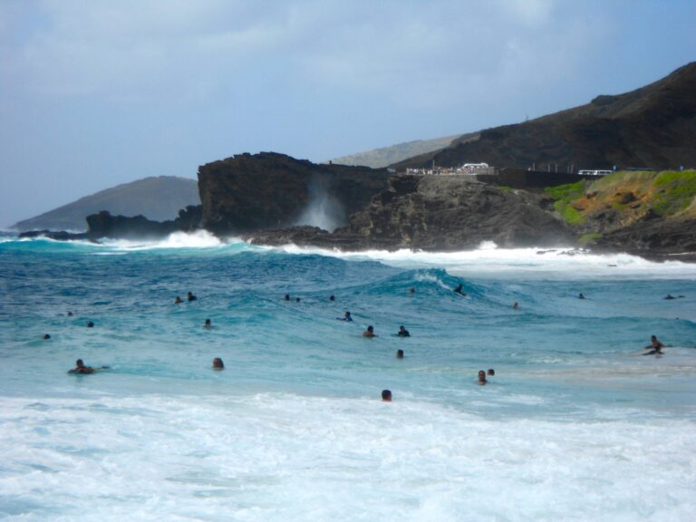 Swimmers and bodyboarders in the surf at Sandy Beach on a windy day with the Hālona Blowhole Lookout behind