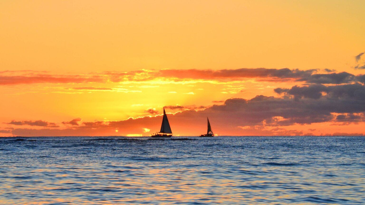 Two sailboats silhouetted against an orange sunset over the ocean off Waikiki