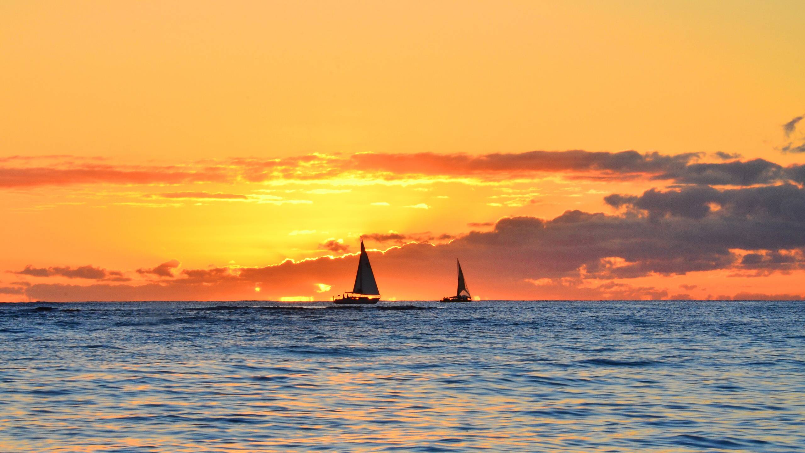 Two sailboats silhouetted against an orange sunset over the ocean off Waikiki